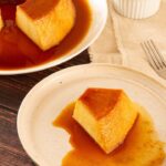 Imagem de uma fatia de pudim em um prato, com um garfo ao lado e outro prato com o restante do pudim ao fundo