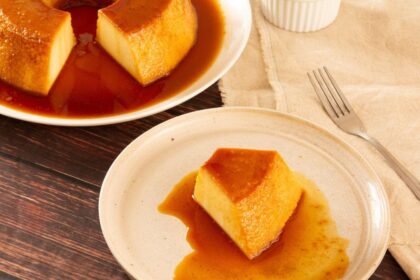 Imagem de uma fatia de pudim em um prato, com um garfo ao lado e outro prato com o restante do pudim ao fundo