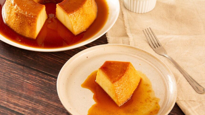 Imagem de uma fatia de pudim em um prato, com um garfo ao lado e outro prato com o restante do pudim ao fundo