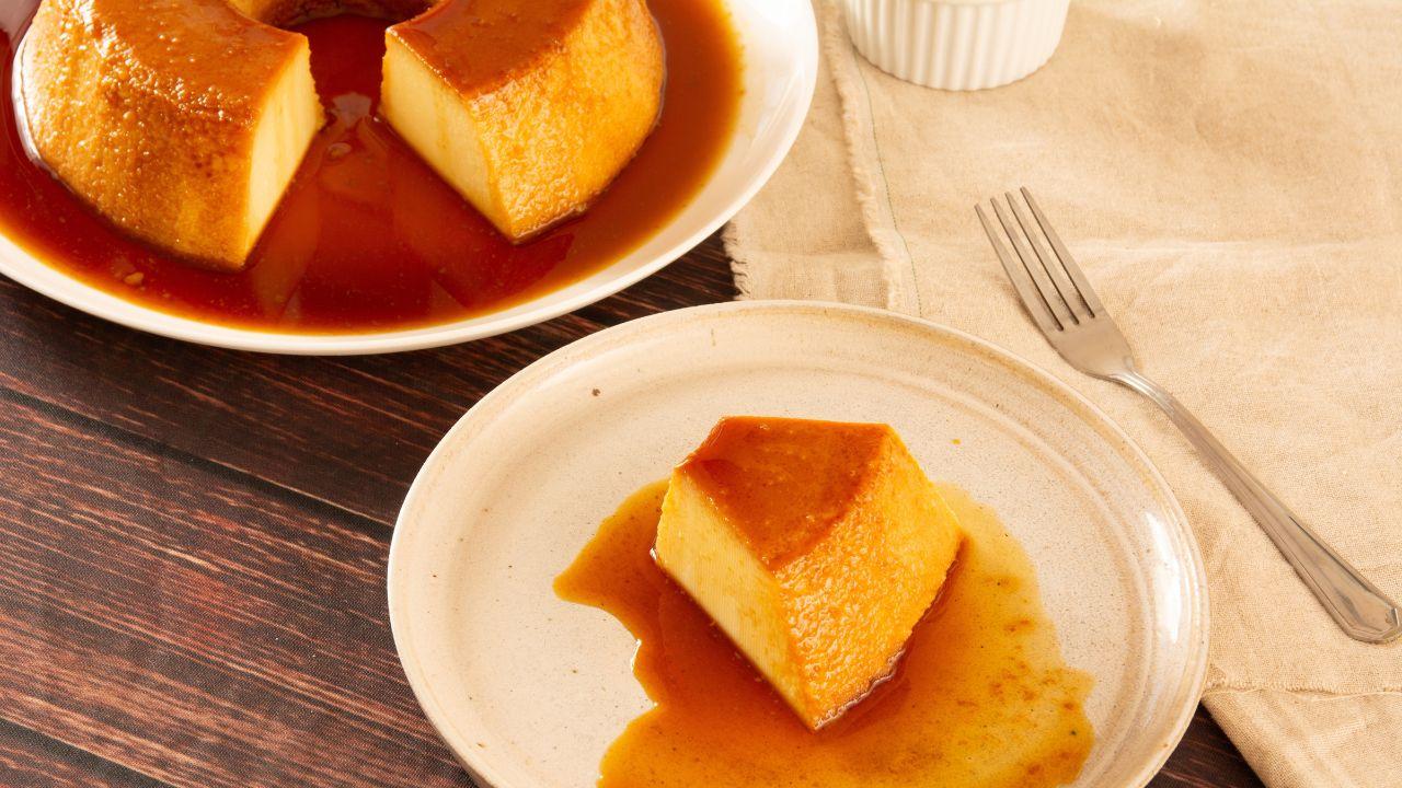 Imagem de uma fatia de pudim em um prato, com um garfo ao lado e outro prato com o restante do pudim ao fundo