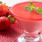 Foto mostra um mousse de morango numa taça de vidro em cima de uma mesa. Ao lado tem morangos in natura.