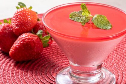 Foto mostra um mousse de morango numa taça de vidro em cima de uma mesa. Ao lado tem morangos in natura.