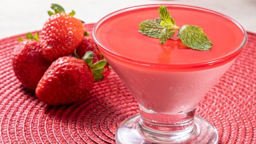 Foto mostra um mousse de morango numa taça de vidro em cima de uma mesa. Ao lado tem morangos in natura.