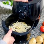 Foto mostra uma mão manuseando a air fryer com batata frita dentro.
