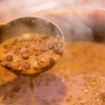 Foto mostra uma panela de feijão cozido quente e uma concha tirando o feijão da panela.