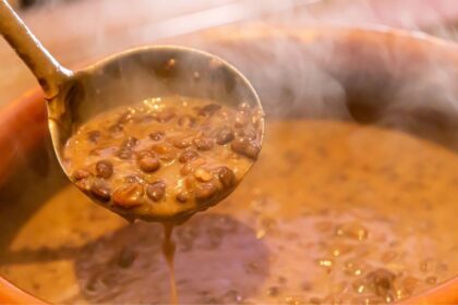 Foto mostra uma panela de feijão cozido quente e uma concha tirando o feijão da panela.