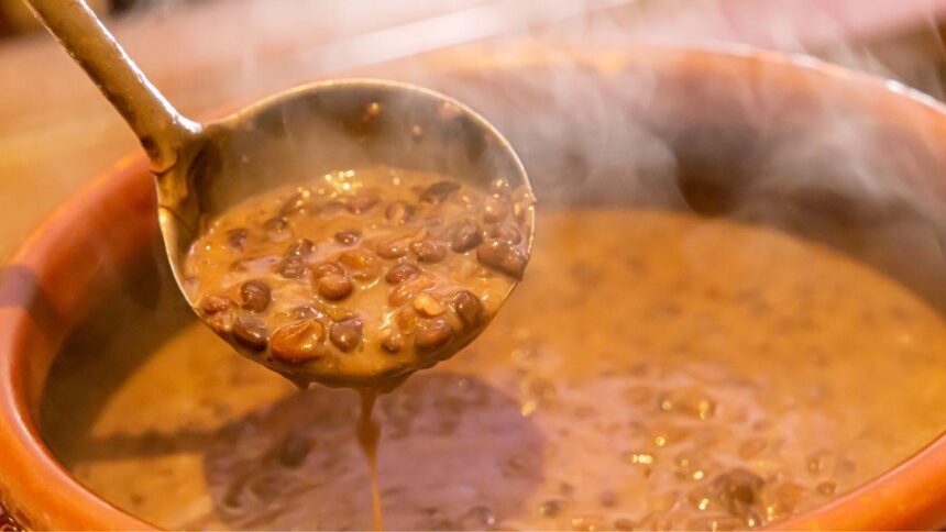 Foto mostra uma panela de feijão cozido quente e uma concha tirando o feijão da panela.