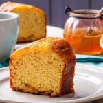 Imagem de uma fatia de bolo de laranja em cima de um prato raso branco; há uma xícara branca, meia laranja e um pote de mel ao fundo