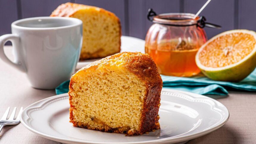 Imagem de uma fatia de bolo de laranja em cima de um prato raso branco; há uma xícara branca, meia laranja e um pote de mel ao fundo