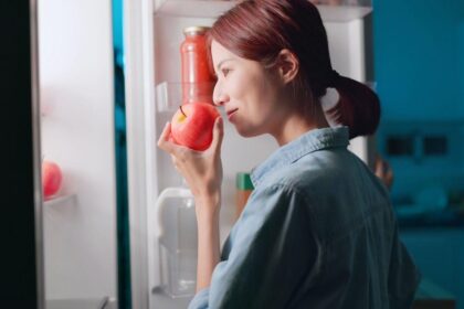 Mulher abrindo a geladeira e segurando uma maçã na mão.