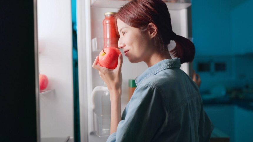 Mulher abrindo a geladeira e segurando uma maçã na mão.