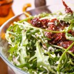 Cumbuca de salada verde com tomate seco em cima de uma mesa.