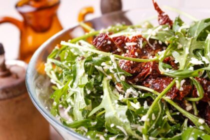 Cumbuca de salada verde com tomate seco em cima de uma mesa.