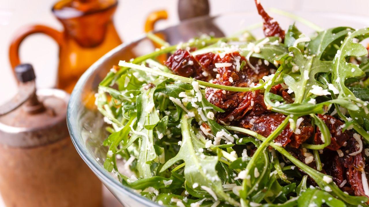 Cumbuca de salada verde com tomate seco em cima de uma mesa.