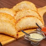 Imagem de uma porção de pastel (6 unidades) em cima de uma tábua de madeira; há também dois potinhos de vidro com molhos na frente