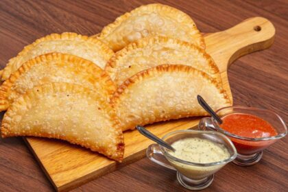 Imagem de uma porção de pastel (6 unidades) em cima de uma tábua de madeira; há também dois potinhos de vidro com molhos na frente