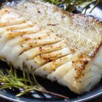Foto mostra um filé de peixe branco suculento em cima de um prato com ramos de alecrim do lado.