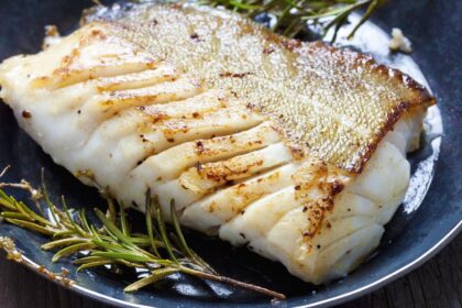 Foto mostra um filé de peixe branco suculento em cima de um prato com ramos de alecrim do lado.