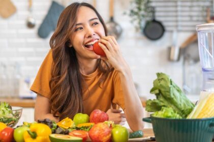 Foto mostra mulher na frente de uma mesa com várias frutas na frente e ela mordendo uma maçã.