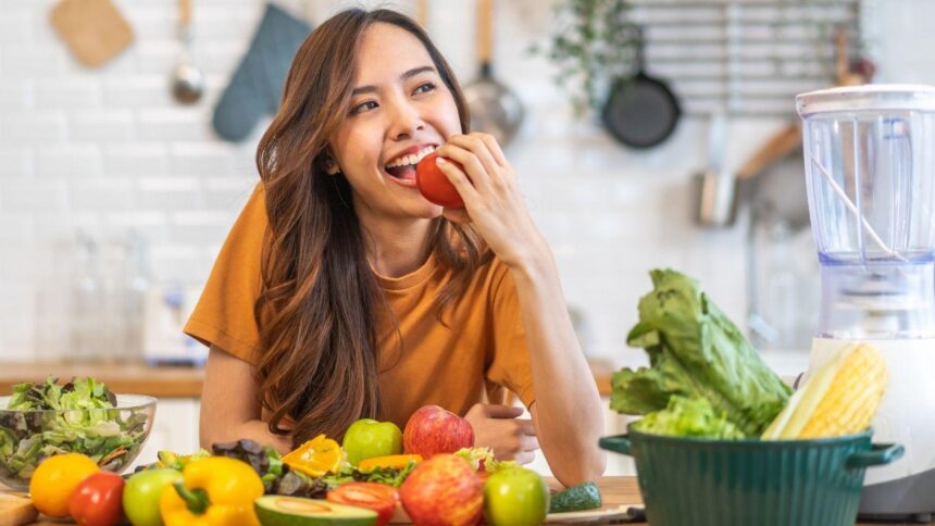 Foto mostra mulher na frente de uma mesa com várias frutas na frente e ela mordendo uma maçã.