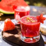 Foto mostra copo de drink de melancia em cima da mesa ao ar livre.