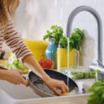 Pessoa lavando panela na pia da cozinha enquanto prepara alimentos frescos, mostrando hábito de limpeza durante o preparo das refeições.