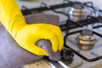 Foto mostra mulher com luva amarela limpando o fogão de alumínio.