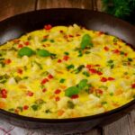Foto mostra um omelete com legumes dentro da frigideira preta em cima da mesa.