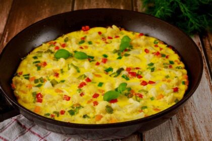 Foto mostra um omelete com legumes dentro da frigideira preta em cima da mesa.