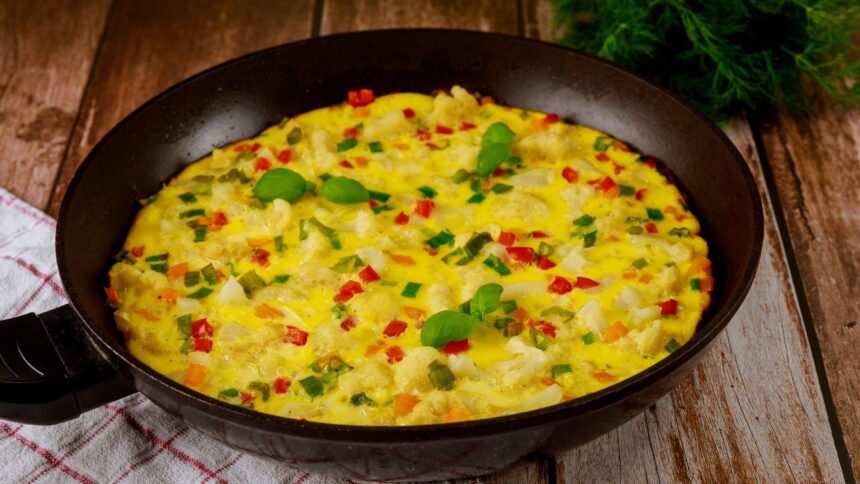 Foto mostra um omelete com legumes dentro da frigideira preta em cima da mesa.