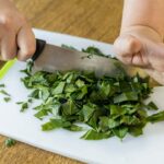Planta verde sendo cortada em pedacinhos numa tábua em cima da mesa.