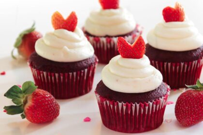 Imagem de quatro cupcakes vermelhos, com cobertura branca e morangos em cima, em um prato