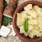 Imagem de mandioca cozida em um pote marrom escuro com raízes de mandioca do lado