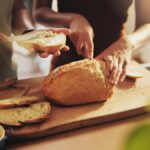 Pão caseiro em cima de uma tábua de madeira com pessoas mexendo e cortando o pão.