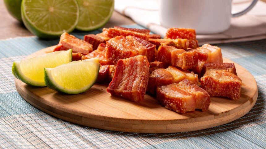 Imagem de pedaços de torresmo em uma tábua de madeira com rodelas de limão ao lado