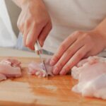 Foto mostra tábua de madeira com frango em cima de uma bancada na cozinha e alguém cortando o frango.
