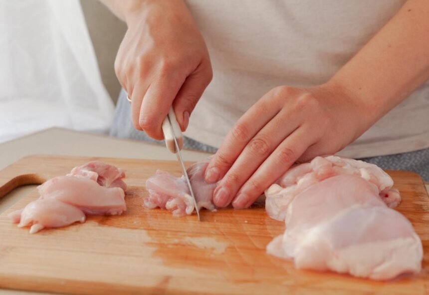 Foto mostra tábua de madeira com frango em cima de uma bancada na cozinha e alguém cortando o frango.
