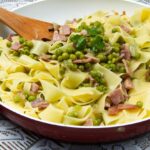 Foto mostra macarrão em espaguete dentro da frigideira com ervilha e outros legumes em cima.