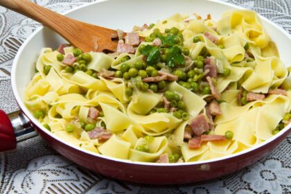 Foto mostra macarrão em espaguete dentro da frigideira com ervilha e outros legumes em cima.