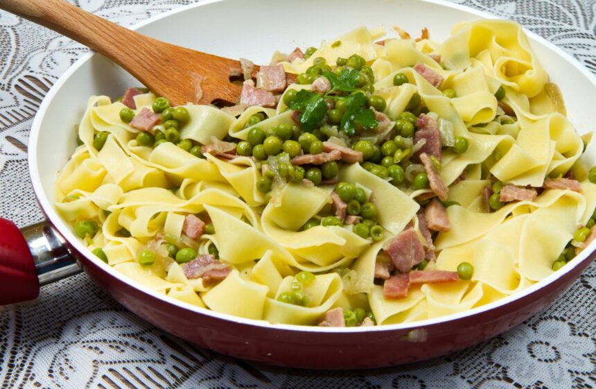 Foto mostra macarrão em espaguete dentro da frigideira com ervilha e outros legumes em cima.