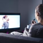 Mulher de costas sentada no sofá enquanto come pipoca e assiste televisão no escuro.