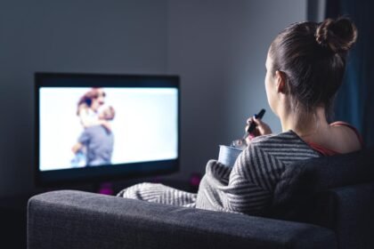 Mulher de costas sentada no sofá enquanto come pipoca e assiste televisão no escuro.