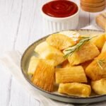 Imagem de uma porção de mandioca frita em um prato com molho vermelho ao fundo