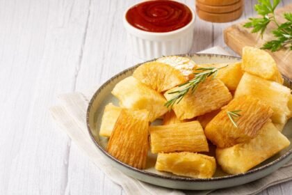 Imagem de uma porção de mandioca frita em um prato com molho vermelho ao fundo