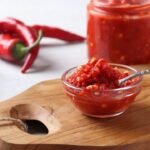 Imagem de um molho de pimenta em um pequeno pote de vidro com pimentas dedo de moça ao redor