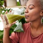 Mulher negra, cabelo crespo loiro e curto, bebendo um copo de suco verde.