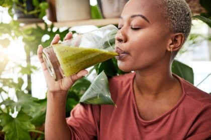 Mulher negra, cabelo crespo loiro e curto, bebendo um copo de suco verde.