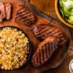 Imagem de um prato com farofa e arroz com carnes de churrasco em volta