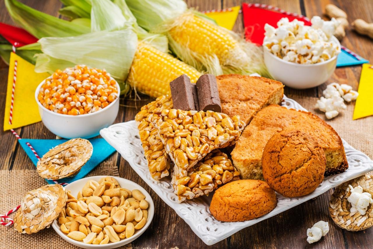 Foto mostra milho, paçoca, amendoim, broa de milho em cima de uma mesa.