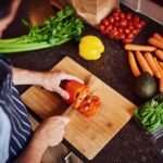 Foto mostra pessoa cortando legumes em uma tábua na bancada da cozinha.